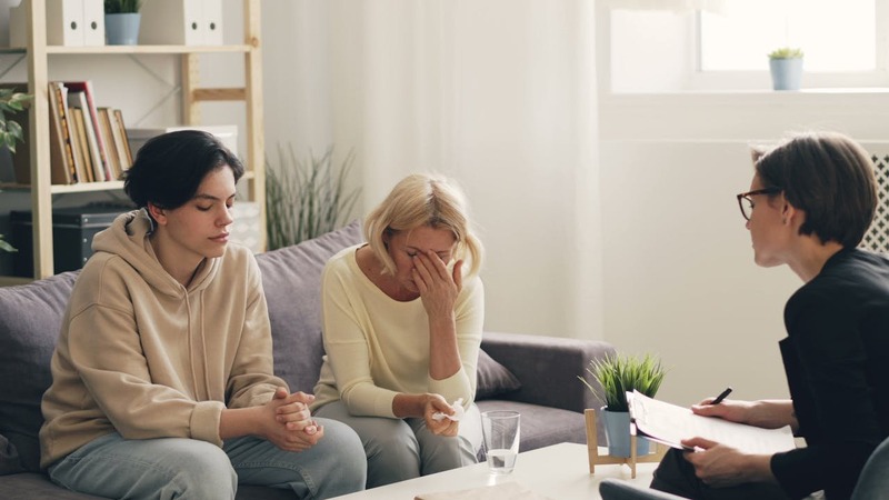 Three people in a room, two on a couch and one on a chair with a clipboard, possibly in a counseling session.
